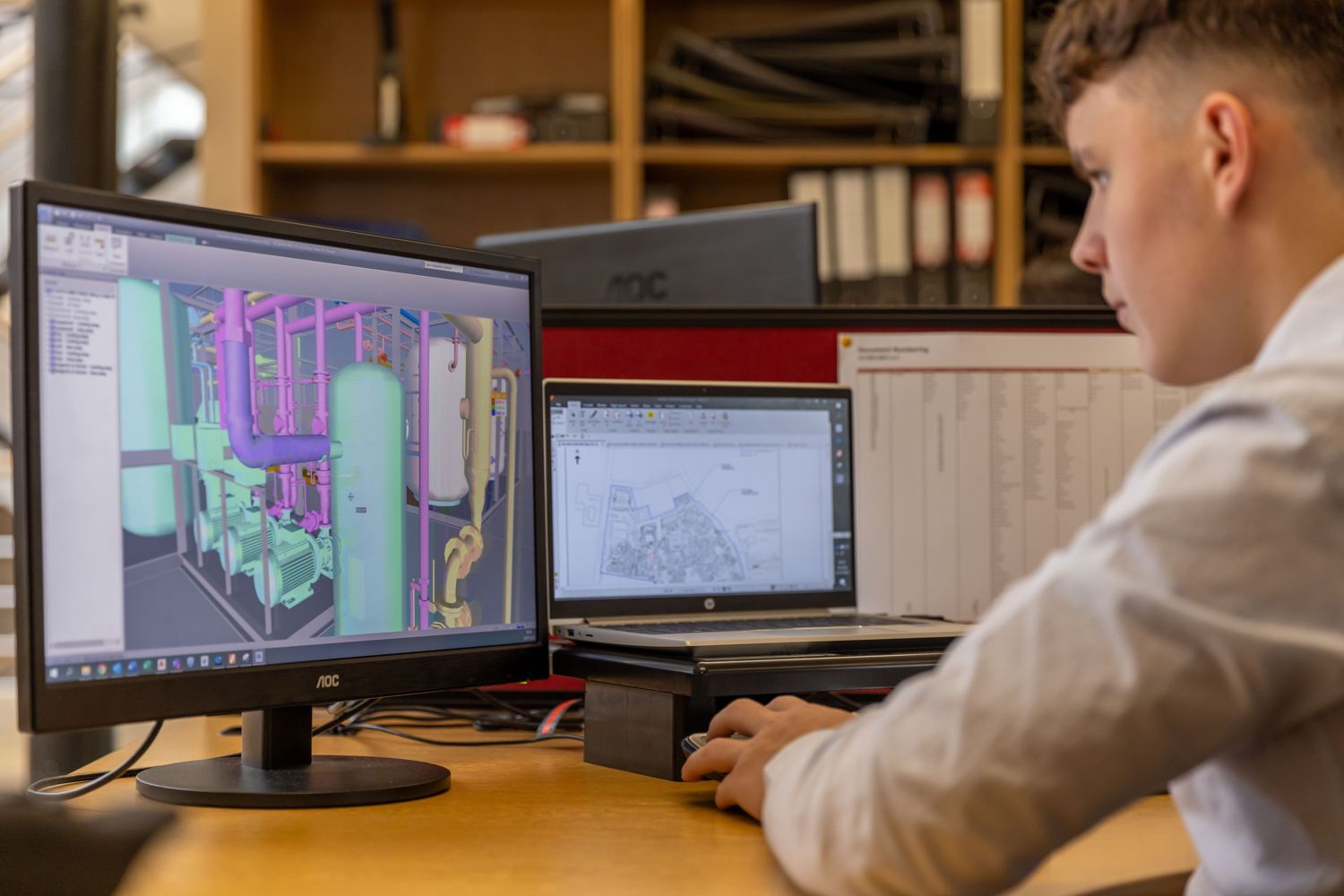 An engineer at a desk with a 3D plant-room model on one screen and a 2D drawing on a laptop