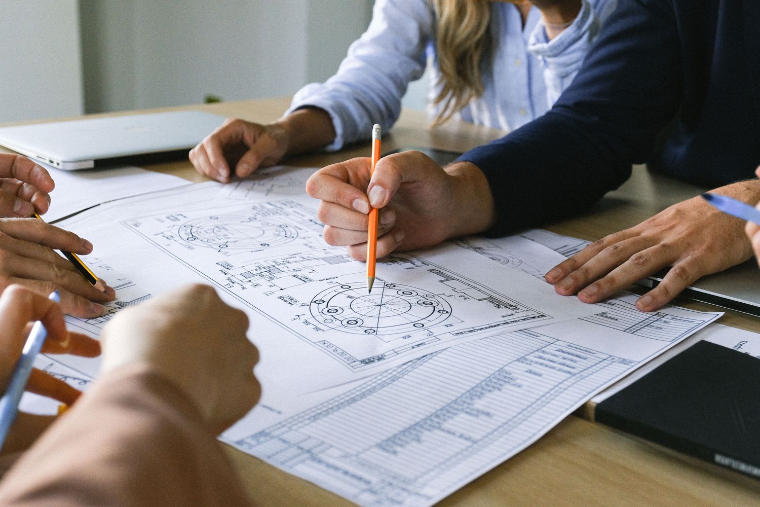 Colleagues around a table reviewing technical drawings with pencils and a laptop